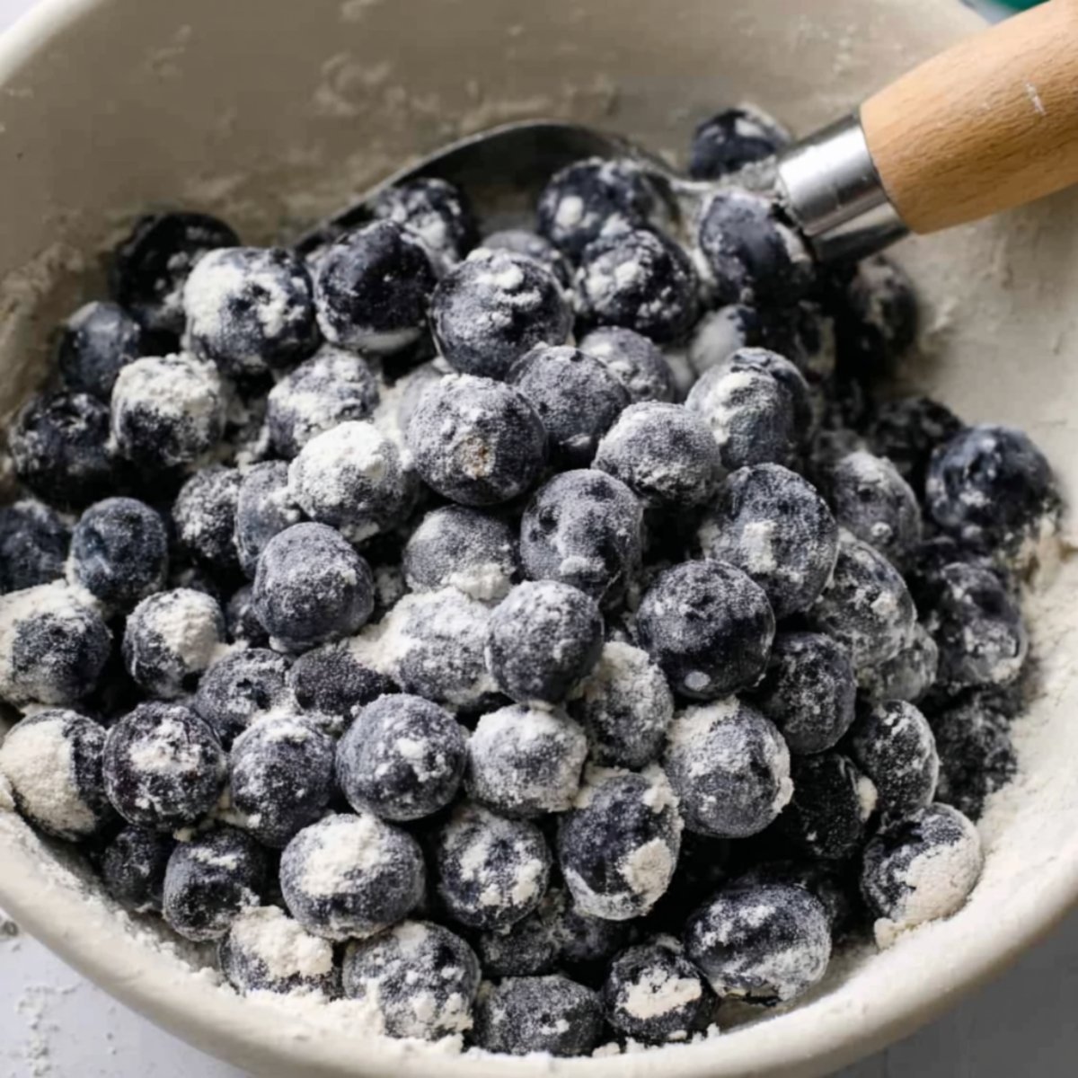 The Best lemon Blueberry Bread Recipe 11 Close up of fresh blueberries coated in flour, ready to be added to a batter. The blueberries are in a white bowl with a spoon resting inside.