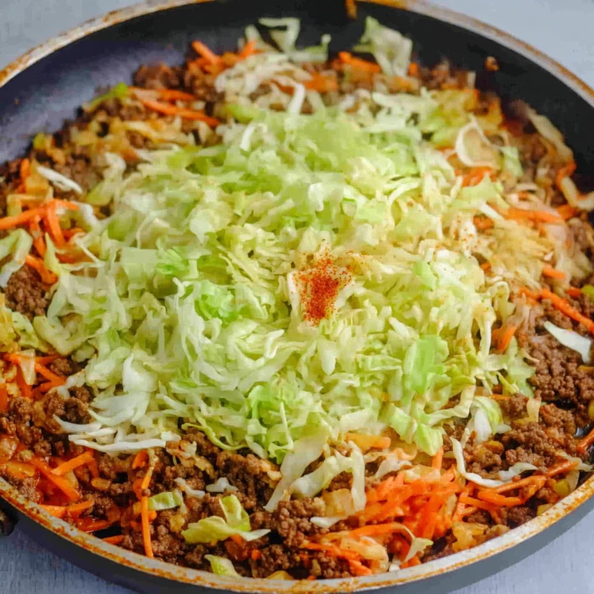 Delicious Egg Roll in a Bowl Recipe 11 A skillet filled with browned ground beef, shredded carrots, and chopped cabbage, with seasonings sprinkled on top.