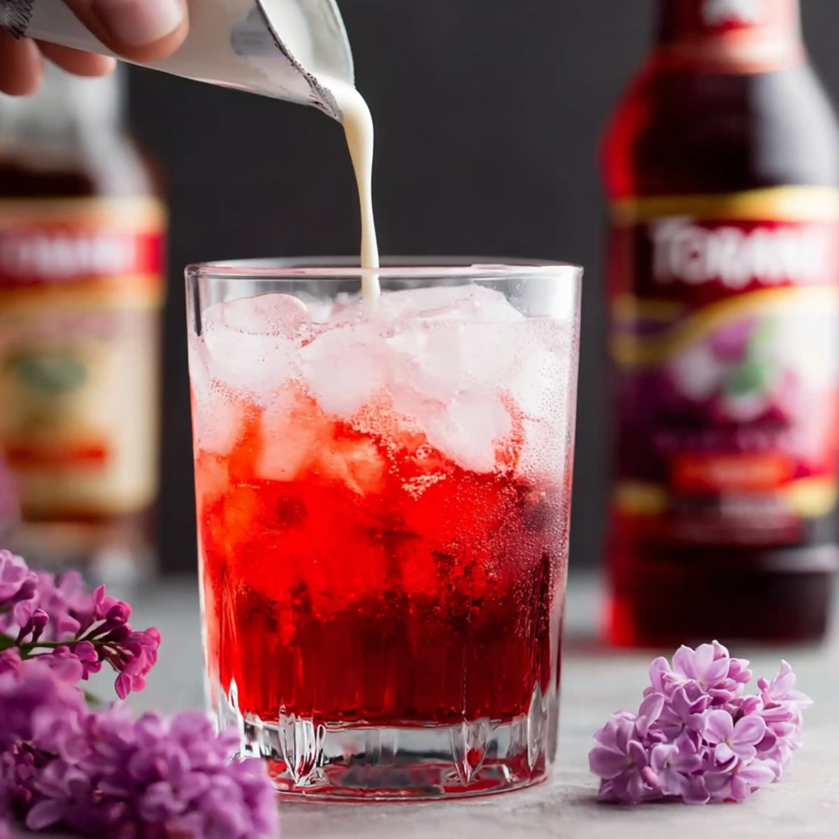 The Best Italian Cream Soda Recipe 11 A hand pouring cream into a glass filled with ice and a red beverage, with a bottle of syrup in the background and purple lilac flowers surrounding the glass.