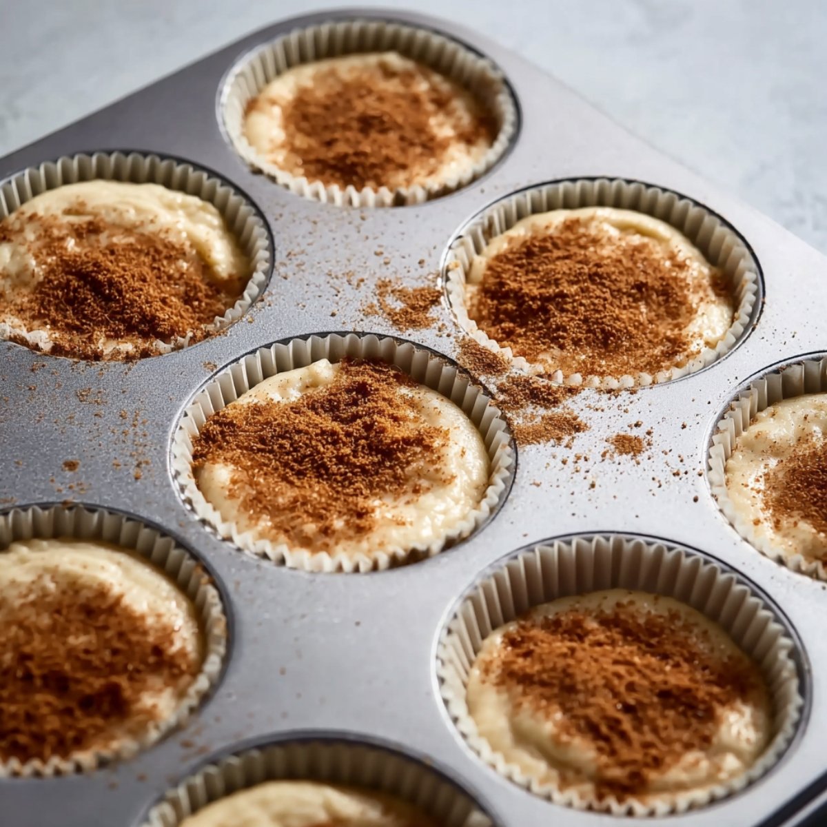 The Best Cinnamon Roll Cupcakes Recipe 11 Close up of a muffin tin filled with cupcake liners, each containing batter topped with a sprinkle of cinnamon sugar. The cinnamon is lightly dusted over the creamy batter, ready to be baked. The scene is illuminated by soft natural lighting, focusing on the muffin tin.