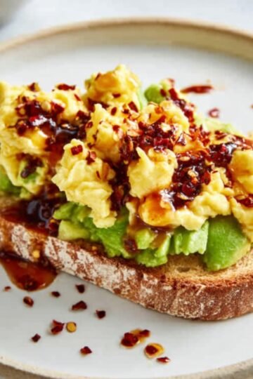 Healthy Avocado Toast Recipe 9 A slice of Avocado Toast bread with mashed avocado spread on top, topped with scrambled eggs, and drizzled with chili oil. The toast is garnished with red chili flakes, creating a vibrant, flavorful breakfast dish.