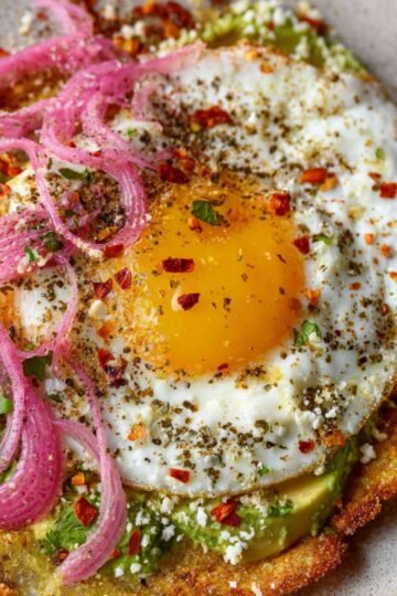 Easy Crispy Feta Fried Eggs Recipe 20 Close-up of a Crispy Feta Fried Eggs with a golden yolk on a tortilla, garnished with pickled red onions, chili flakes, cilantro, and crumbled feta, placed on a bed of creamy avocado.