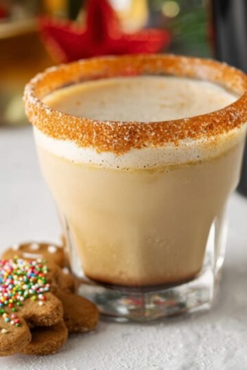 Close up of a Gingerbread White Russian-flavored white Russian in a glass, with a cinnamon-sugar rim. The creamy, frothy beverage is topped with a light foam, and a gingerbread cookie rests beside the glass, complete with colorful sprinkles. The festive, cozy atmosphere is enhanced by a holiday-themed backdrop.