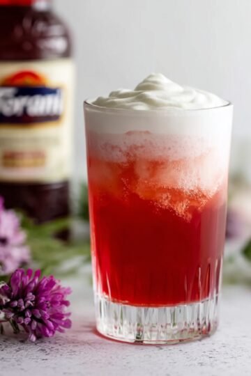 The Best Italian Cream Soda Recipe 7 Italian Cream Soda A tall glass filled with a red drink and topped with a layer of creamy foam, with purple lilac flowers in the background and a bottle of Torani syrup behind.