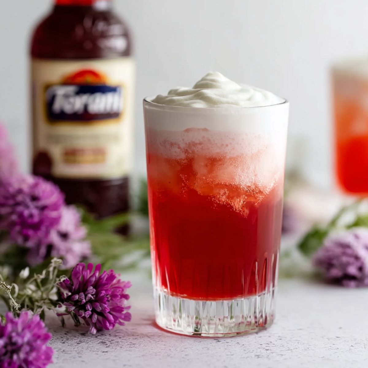 Italian Cream Soda A tall glass filled with a red drink and topped with a layer of creamy foam, with purple lilac flowers in the background and a bottle of Torani syrup behind.