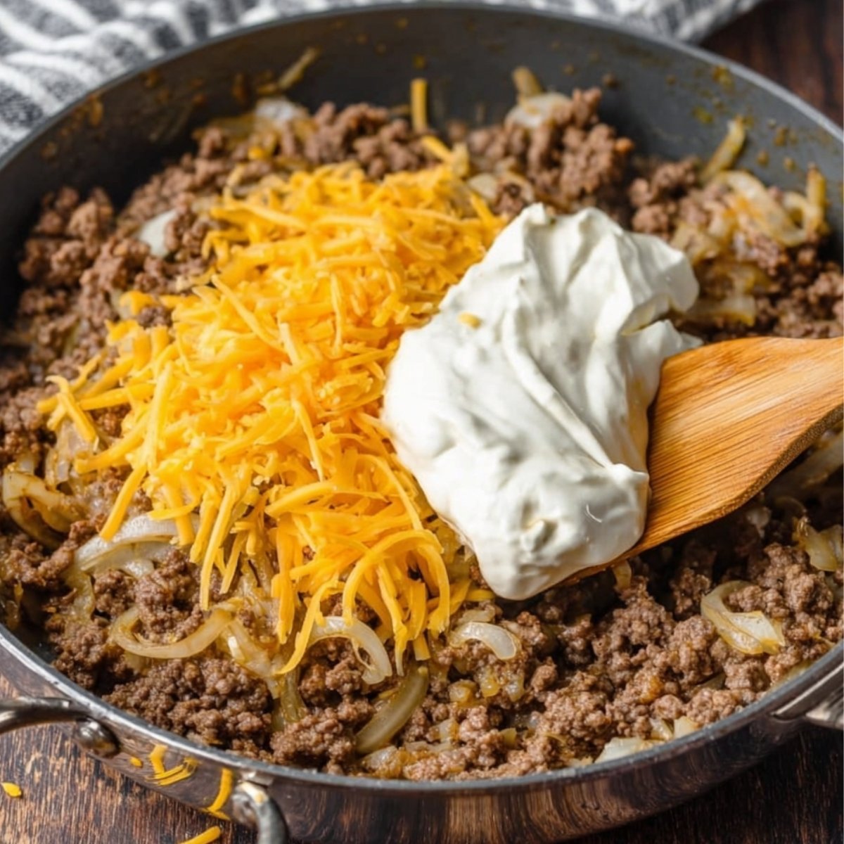Easy Hobo Casserole Recipe 10 A skillet with ground beef, onions, and shredded cheddar cheese, topped with a dollop of sour cream, being stirred with a wooden spoon.