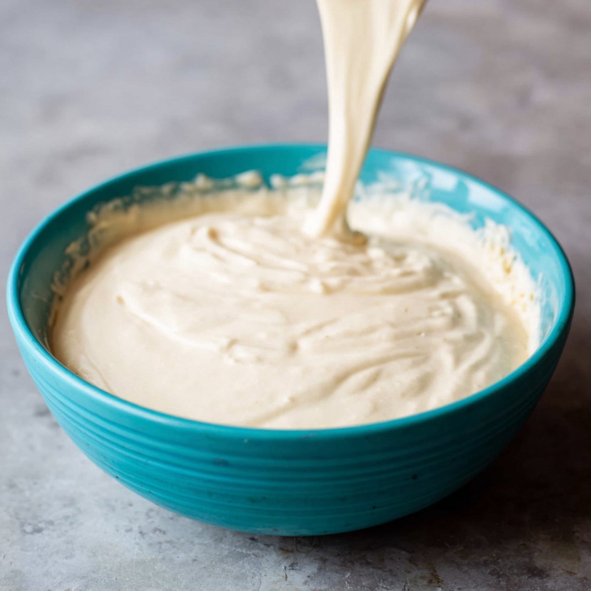 Easy Raspberry Lemonade Cheesecake Recipe 11 Close up of creamy cheesecake filling being poured into a turquoise bowl. The smooth, thick texture of the mixture indicates the start of making a delicious cheesecake.