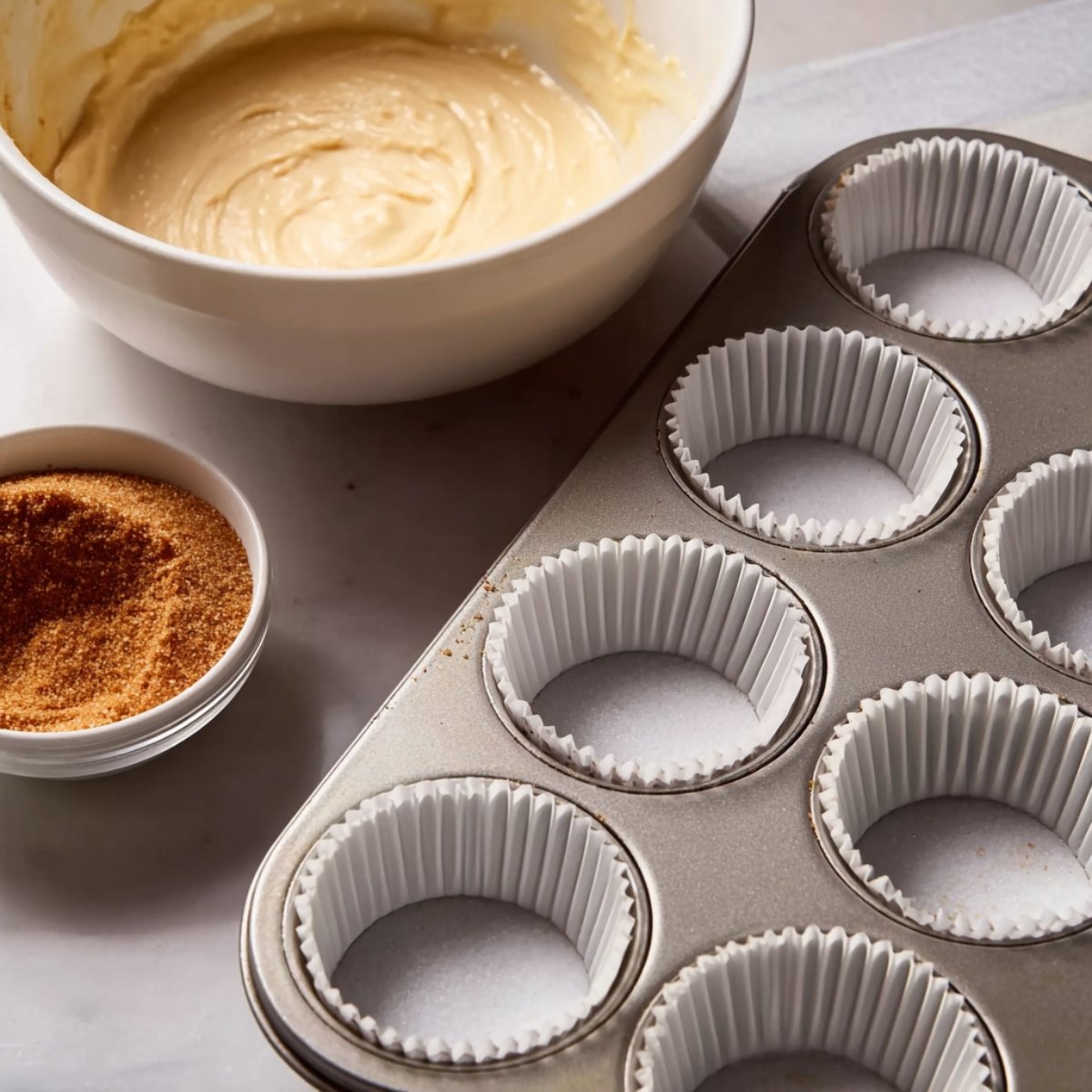 The Best Cinnamon Roll Cupcakes Recipe 10 A bowl of smooth cupcake batter sits next to a muffin tin with empty cupcake liners. A bowl of cinnamon sugar is visible, showing the ingredients for cinnamon roll cupcakes being prepped. The scene captures the preparation process in a clean, minimalist style.