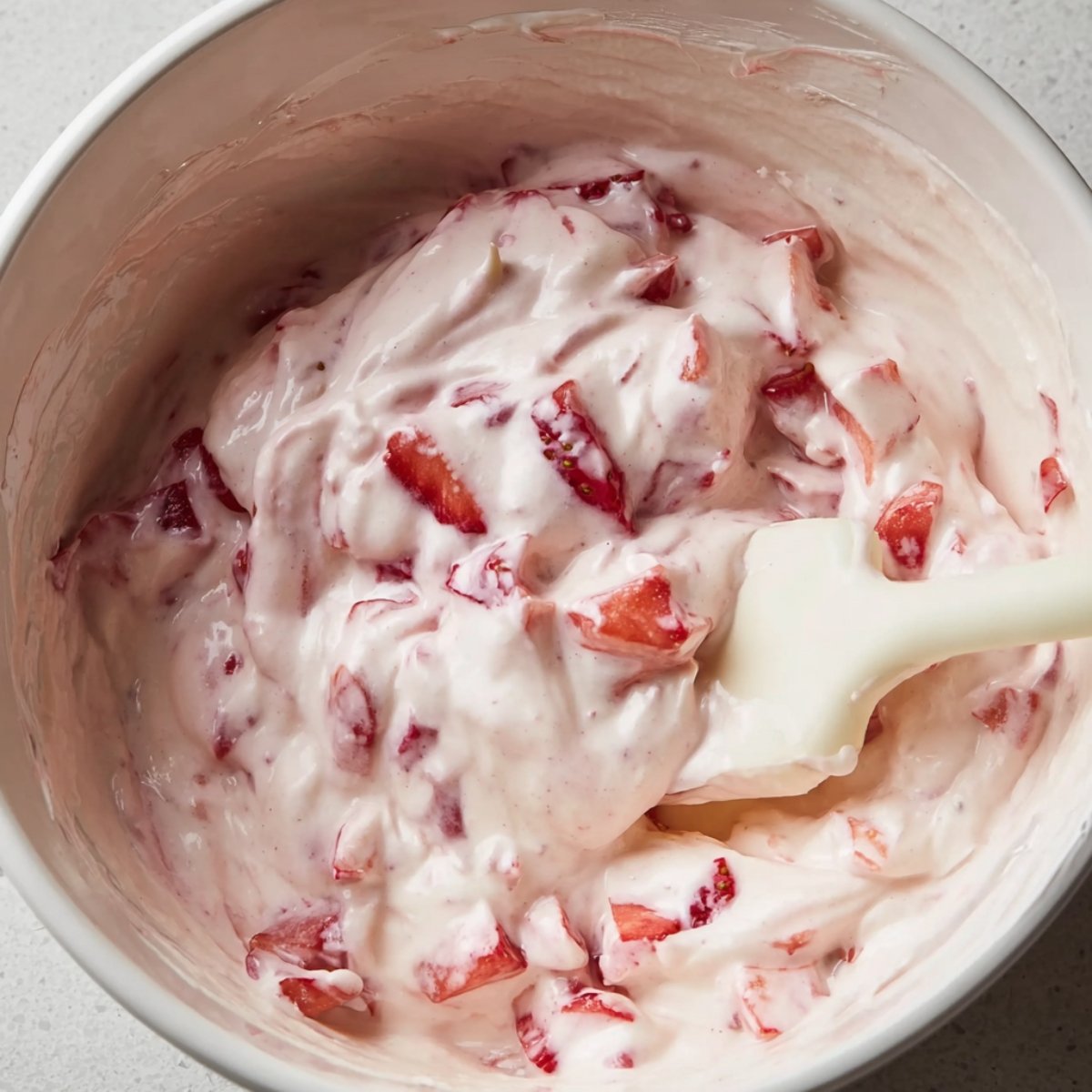 Healthy Chocolate Strawberry Yogurt Clusters 10 Close up of a mixing bowl with chopped strawberries and yogurt blended together. The mixture is creamy with small pieces of fresh strawberries visible, being stirred with a spatula.