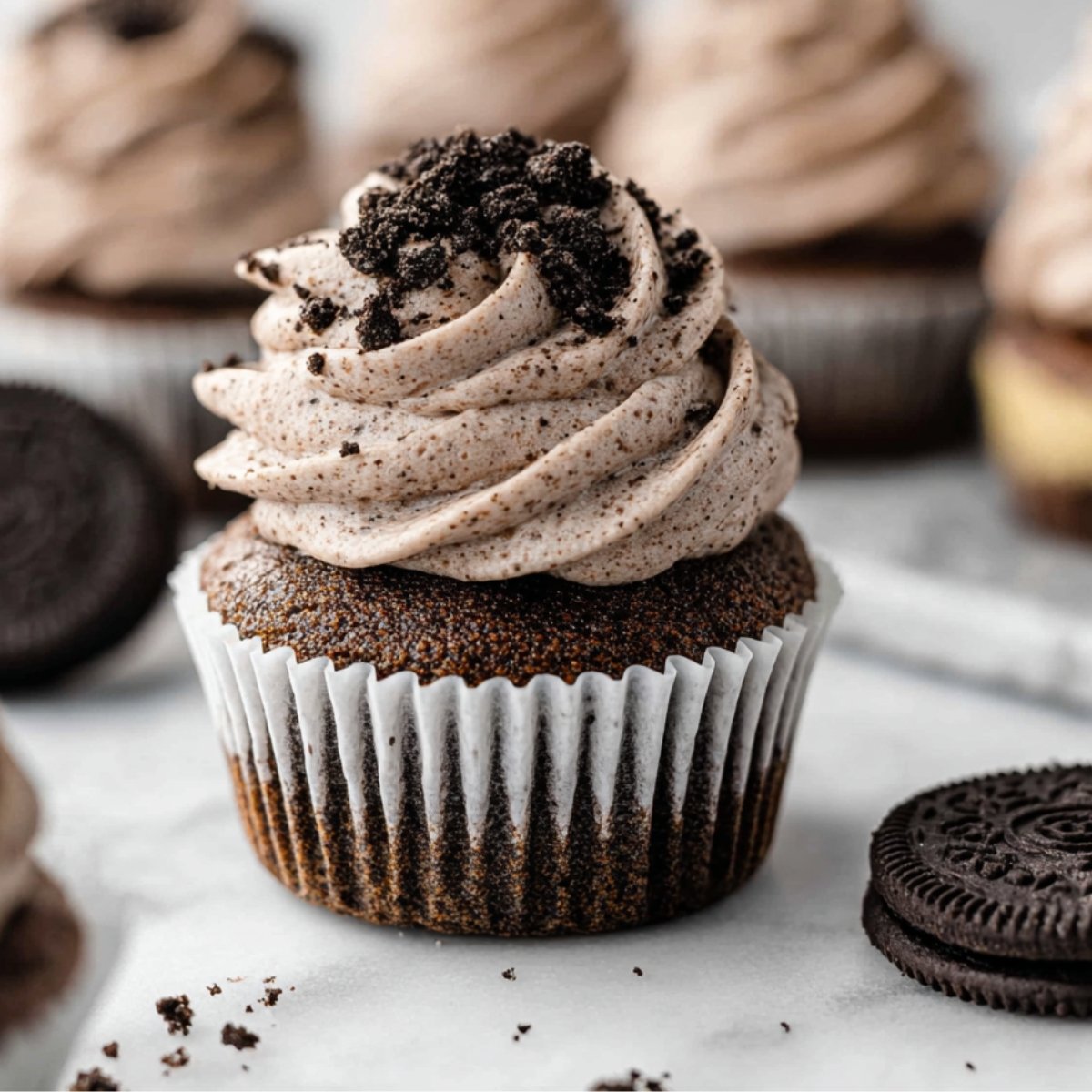 A freshly made cupcake with Oreo Cupcakes frosting, topped with crumbled Oreo cookie pieces, placed on a white background with other cupcakes in the background.