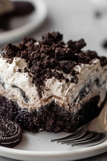 Easy Oreo Poke Cake Recipe 10 Close-up of a slice of Oreo poke cake on a white plate. The cake features layers of dark chocolate, creamy frosting, and crushed Oreos sprinkled on top. An Oreo cookie is placed beside the cake, and a fork is ready to take a bite.
