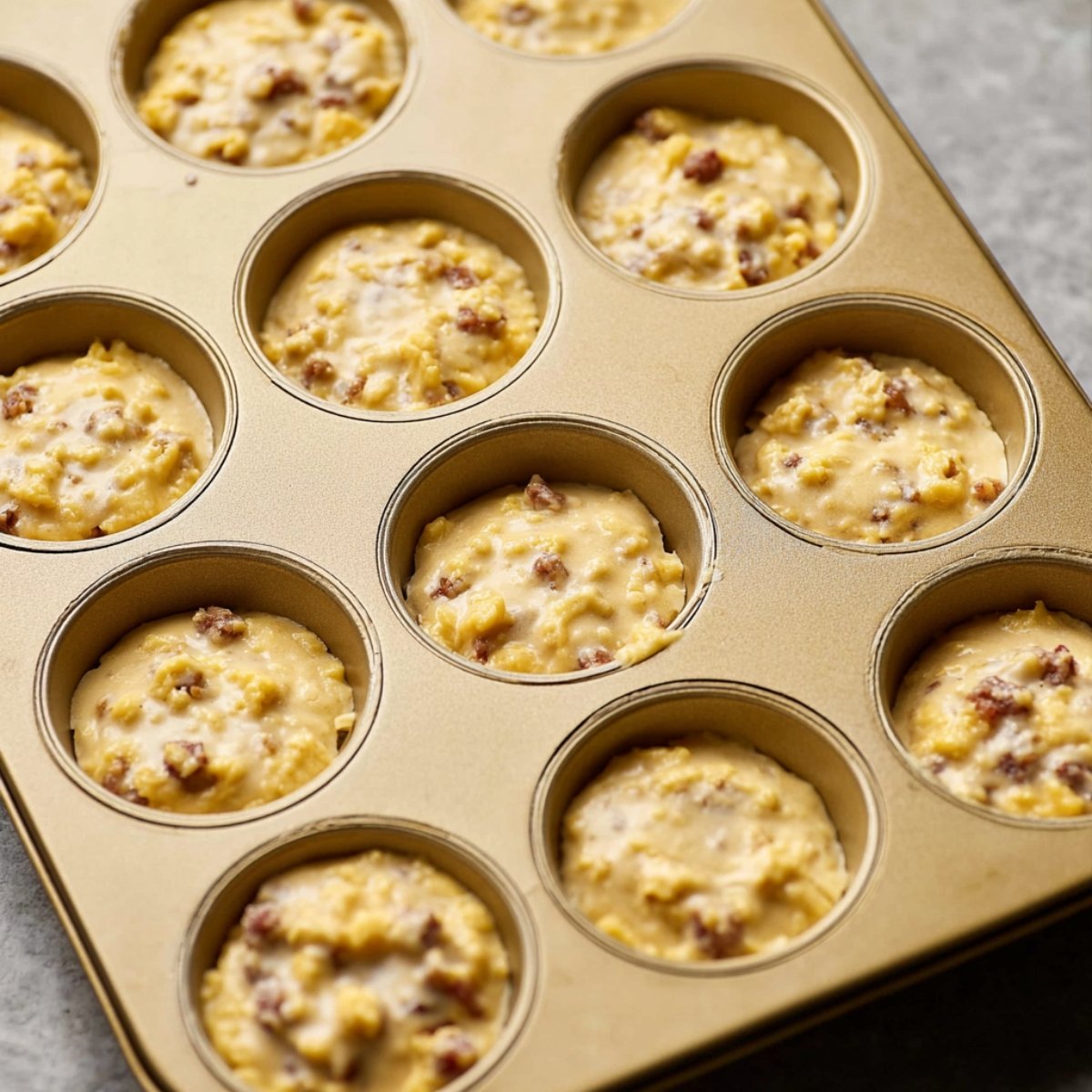 Easy Homemade McGriddle Muffins Recipe 11 Close up of muffin batter with sausage and cheese, placed in a muffin tin. The batter is evenly distributed across the muffin cups, ready to be baked into golden McGriddle muffins.