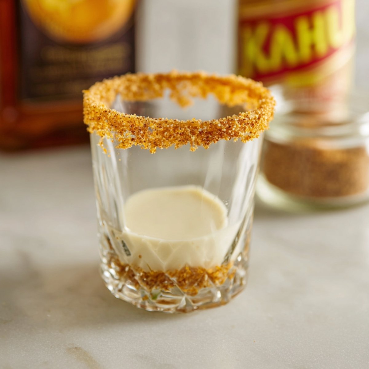 The Best Gingerbread White Russian Recipe 11 Close up of a glass with a cinnamon-sugar rim, with a small amount of creamy liquid visible inside. The rim is coated with a golden-brown mixture of cinnamon and sugar, suggesting the preparation of a festive gingerbread-inspired cocktail. In the background, bottles of Kahlúa and cinnamon powder are partially visible, ready for use.