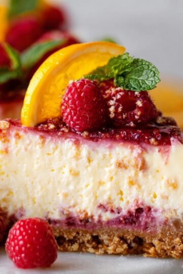 Easy Raspberry Lemonade Cheesecake Recipe 17 Close up of a slice of raspberry lemonade cheesecake topped with fresh raspberries, orange slices, and mint leaves. The smooth, creamy texture of the cheesecake contrasts beautifully with the colorful toppings.