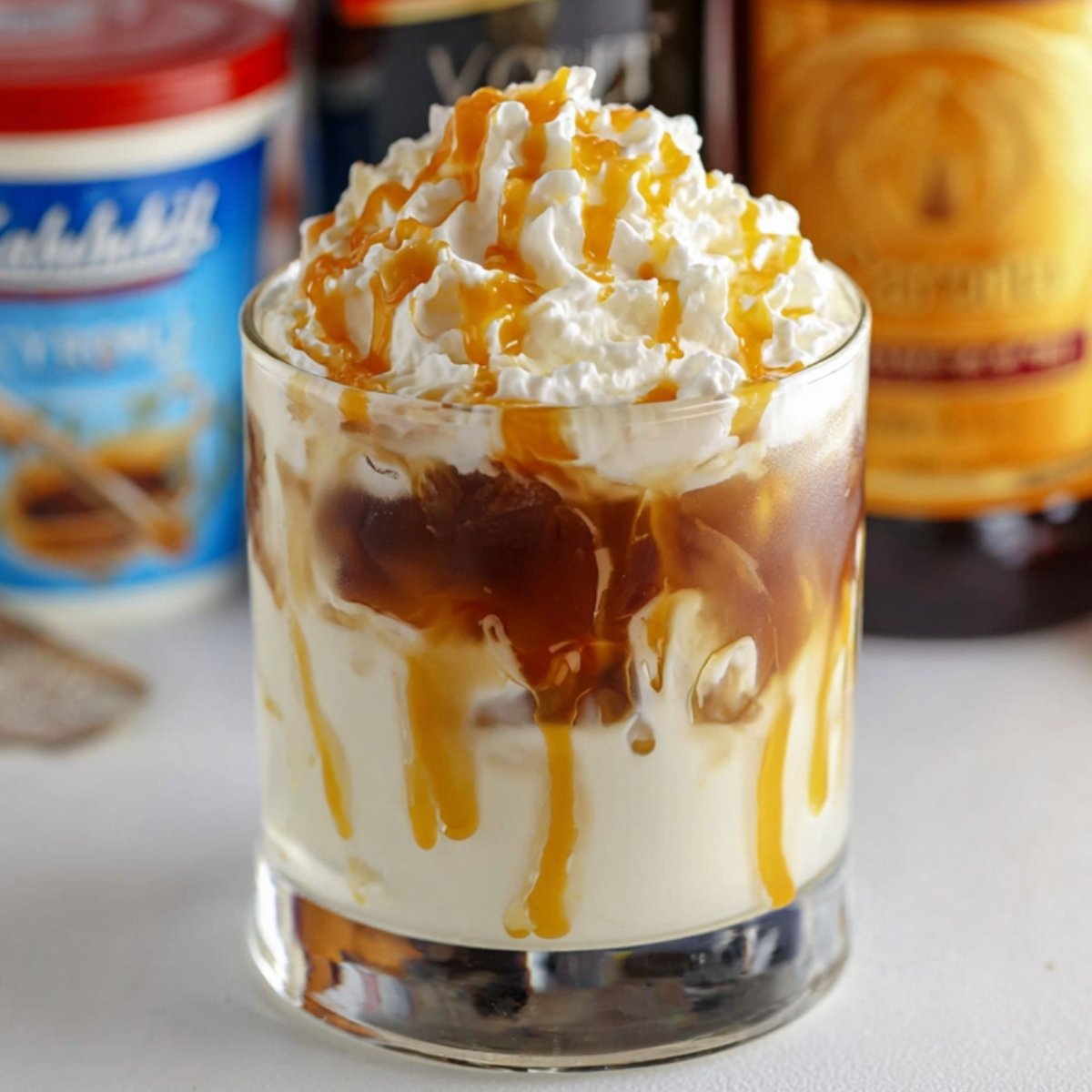 A Salted Caramel White Russian Cocktail in a glass, topped with a generous swirl of whipped cream and drizzled with caramel syrup. The drink has layers of dark coffee and creamy beige, with the caramel syrup running down the sides, creating an inviting and indulgent visual. The background includes some of the cocktail ingredients, such as vodka and Kahlúa.