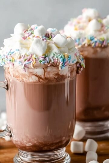Easy Whipped Hot Chocolate Recipe 15 A pair of glass mugs filled with Whipped Hot Chocolate, topped with whipped cream, marshmallows, and rainbow sprinkles. The mugs rest on a wooden tray, with marshmallows scattered around, adding a cozy, indulgent touch to the scene.