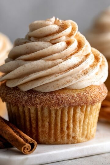 The Best Cinnamon Roll Cupcakes Recipe 6 Close-up of a perfectly frosted cinnamon roll cupcakes. The cupcake is coated in a soft cinnamon swirl frosting, sprinkled with a dusting of cinnamon sugar on top. Cinnamon sticks lie next to the cupcake on a marble tray, completing the cozy, indulgent aesthetic.