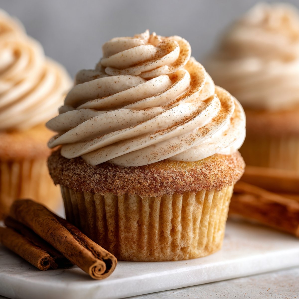 Close-up of a perfectly frosted cinnamon roll cupcakes. The cupcake is coated in a soft cinnamon swirl frosting, sprinkled with a dusting of cinnamon sugar on top. Cinnamon sticks lie next to the cupcake on a marble tray, completing the cozy, indulgent aesthetic.