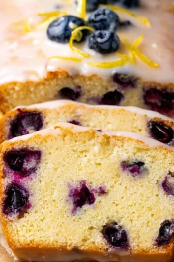 The Best lemon Blueberry Bread Recipe 21 Close up of a freshly baked lemon blueberry bread loaf cake drizzled with a glaze, garnished with fresh blueberries and lemon zest, with slices showing the blueberry-filled interior.