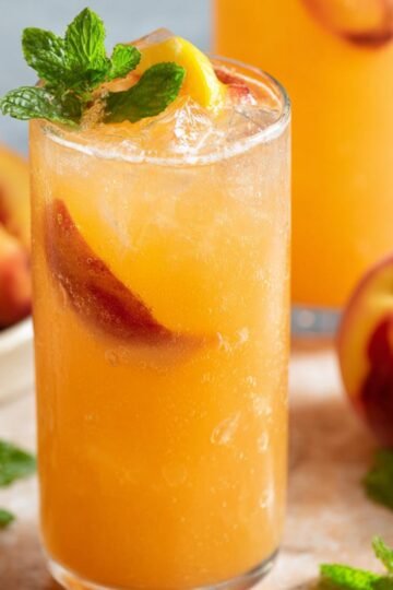 Delicious Peach Lemonade Recipe 16 A close-up of a glass filled with peach lemonade, ice cubes, and slices of peach, garnished with fresh mint. In the background, a whole peach and a few mint leaves are visible, adding to the refreshing vibe.