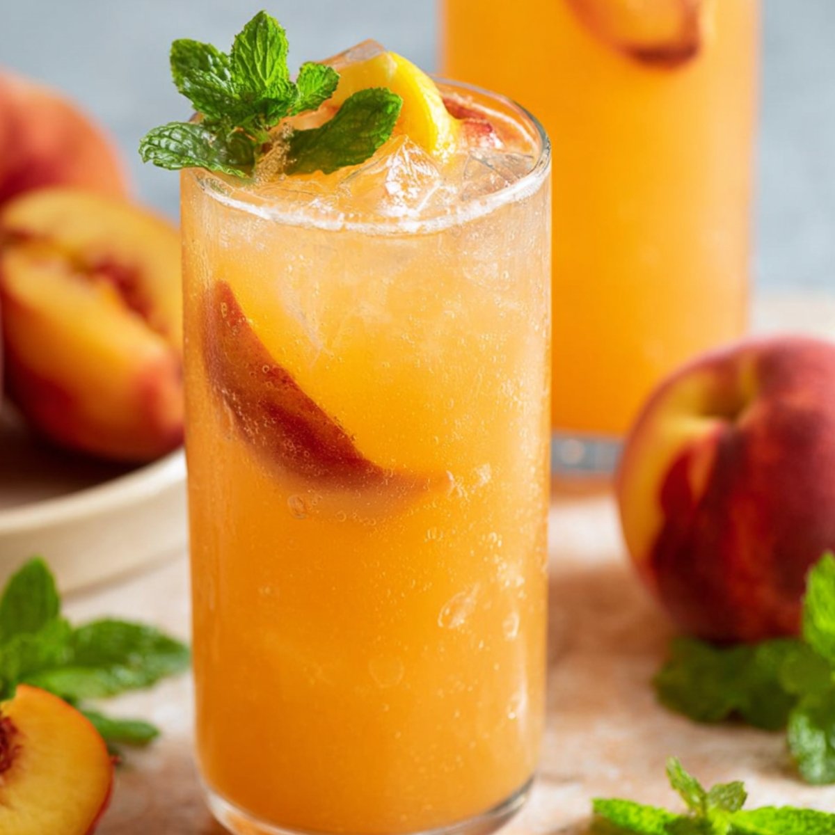 A close-up of a glass filled with peach lemonade, ice cubes, and slices of peach, garnished with fresh mint. In the background, a whole peach and a few mint leaves are visible, adding to the refreshing vibe.