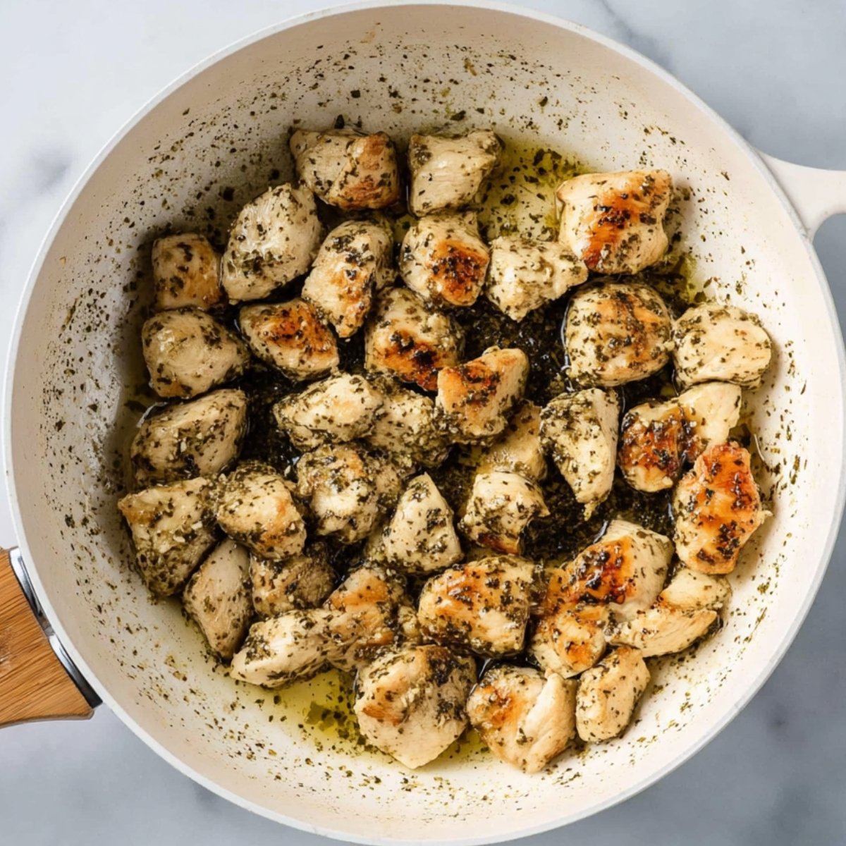 Best Creamy Chicken Pesto Pasta 11 The image shows seasoned chicken chunks cooking in a pan with herbs and olive oil.