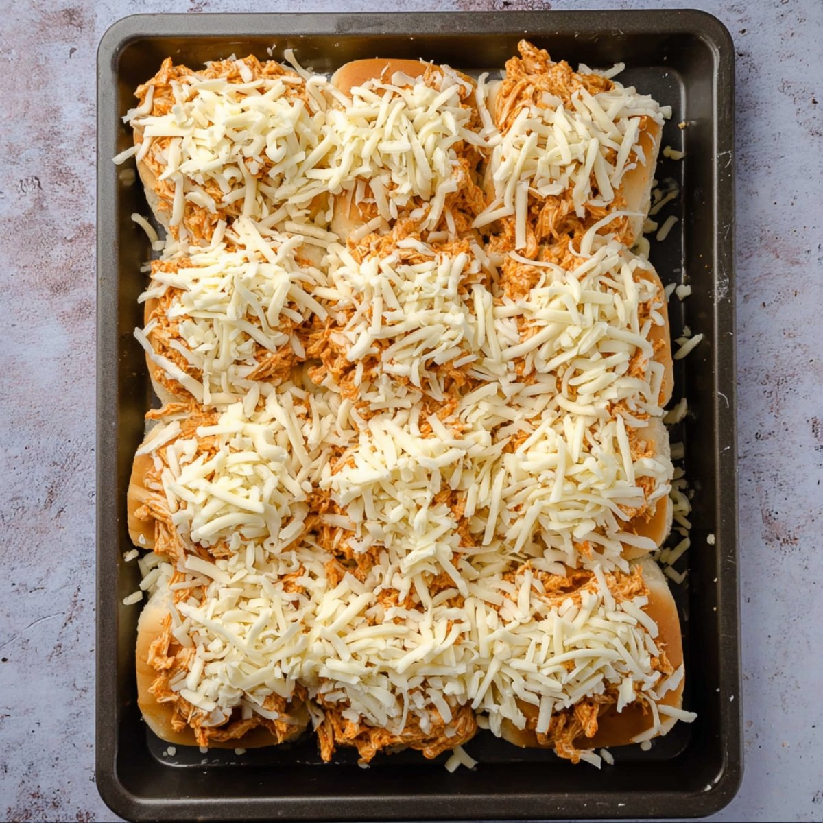 Easy Buffalo Chicken Sliders with Hawaiian Rolls Recipe 11 A baking tray filled with sliders topped with shredded buffalo chicken and a generous layer of shredded mozzarella cheese.