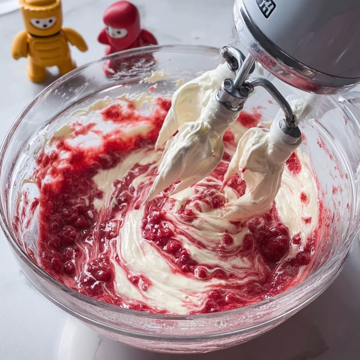 Best Chocolate Strawberry Cake Recipe 11 Whisking raspberry buttercream frosting with a hand mixer, with two figurines in the background.