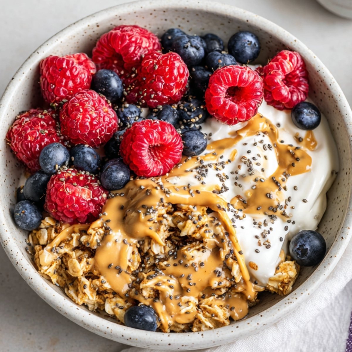 The image shows a bowl Scrambled Oats of oatmeal topped with yogurt, peanut butter, chia seeds, blueberries, and raspberries.
