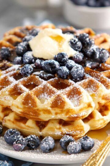 Best Banana Blueberry Waffles 19 A stack of banana Blueberry waffles topped with blueberries, powdered sugar, and a pat of butter.